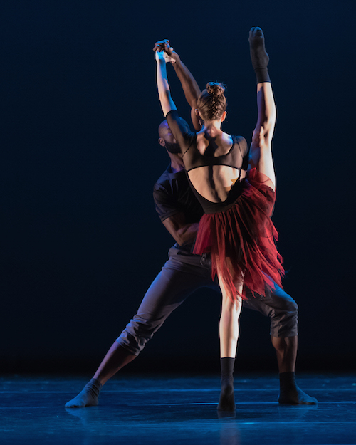 A women extends her right leg in a high developpe with her back turned to the audience. She wears a deep red skirt, intricate black leotard and black calf length socks. Her male counterpart helps her balance by grasping her hands.